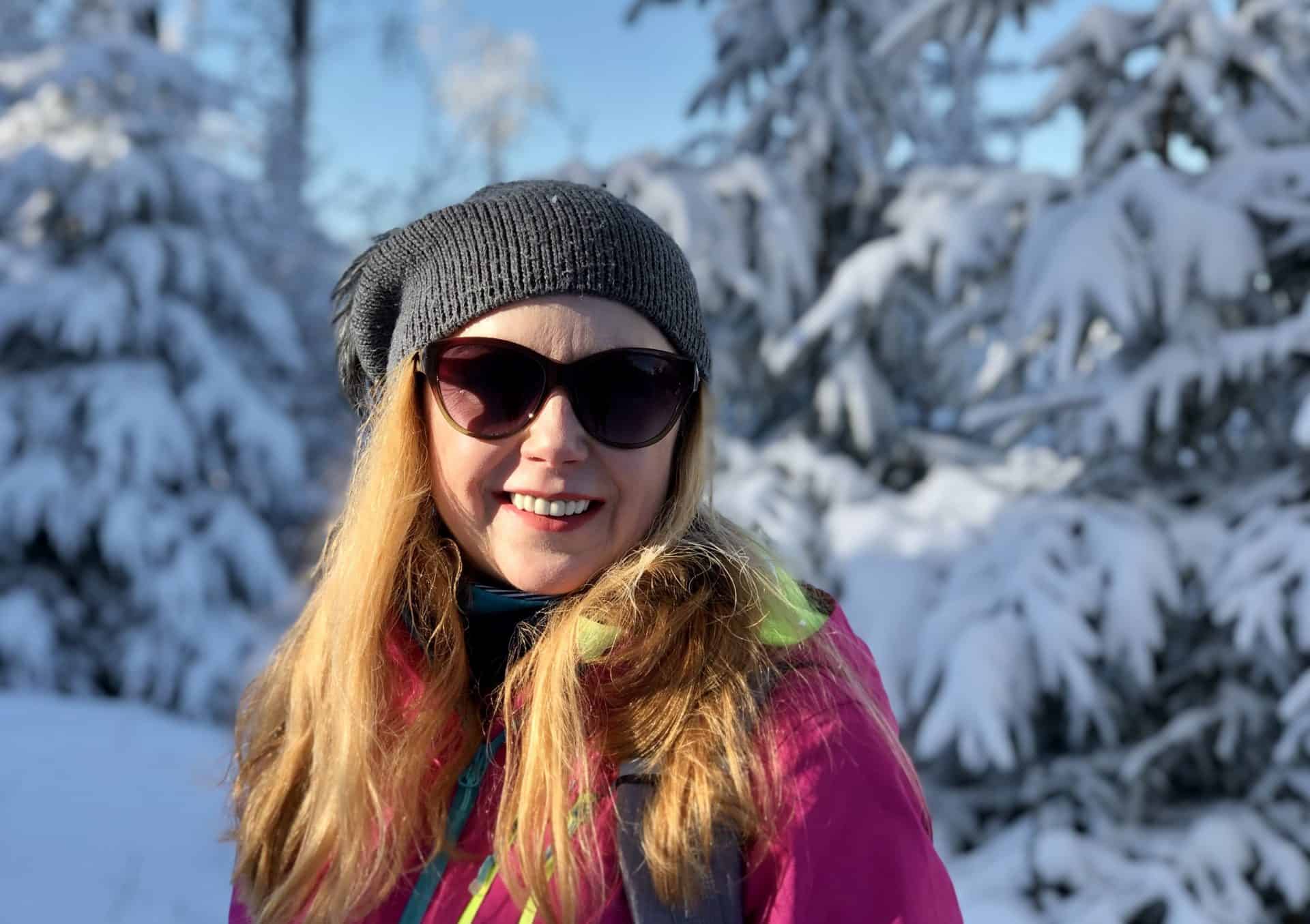 kobieta uśmiechnięta w okularach przeciwsłonecznych i czapce na tle zaśnieżonych drzew zima